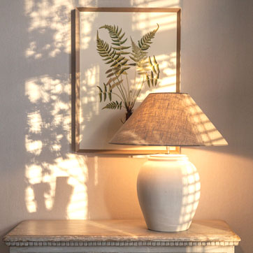 Elegante Keramik Tischlampe vor gerahmter Illustration im warmen Maison Stil