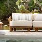 Preview: Tine K Teak Seitenmodul kombiniert mit Mittelelement am Pool — cremefarbene Kissen mit Streifenkissen, recyceltes Teak, mediterrane Gartenkulisse