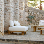 Preview: Tine K Teak Modul Sofa als Einzelsessel auf Kopfsteinpflaster — cremefarbene Kissen, recyceltes Teak, mediterraner Innenhof mit Steinmauer