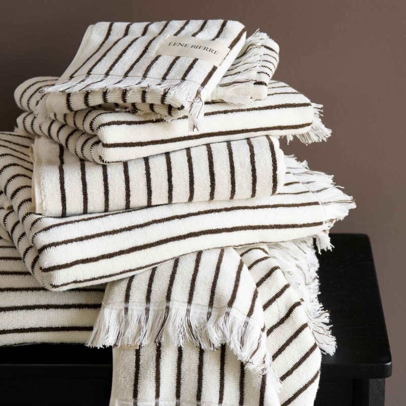 Lene Bjerre Ayana Stripe Handtücher offwhite mit braunen Streifen und Fransen, gestapelt auf dunklem Holztisch