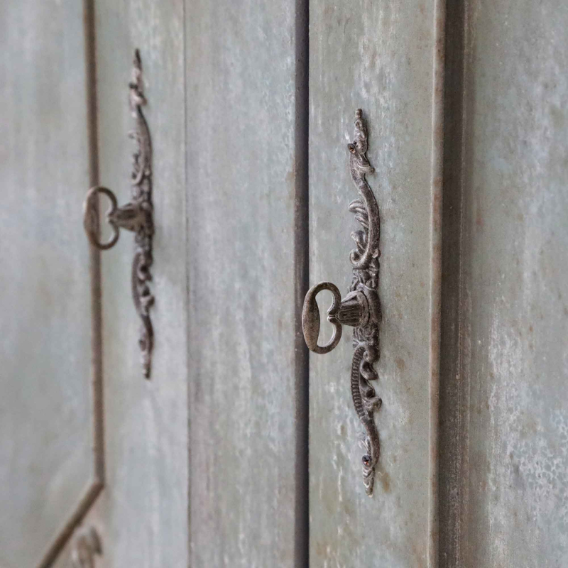 Detailaufnahme provençalischer Schranktisch — ornamentaler Metallbeschlag mit Schlüsselloch auf grau-blauer Kiefernholzoberfläche, antique opal Finish
