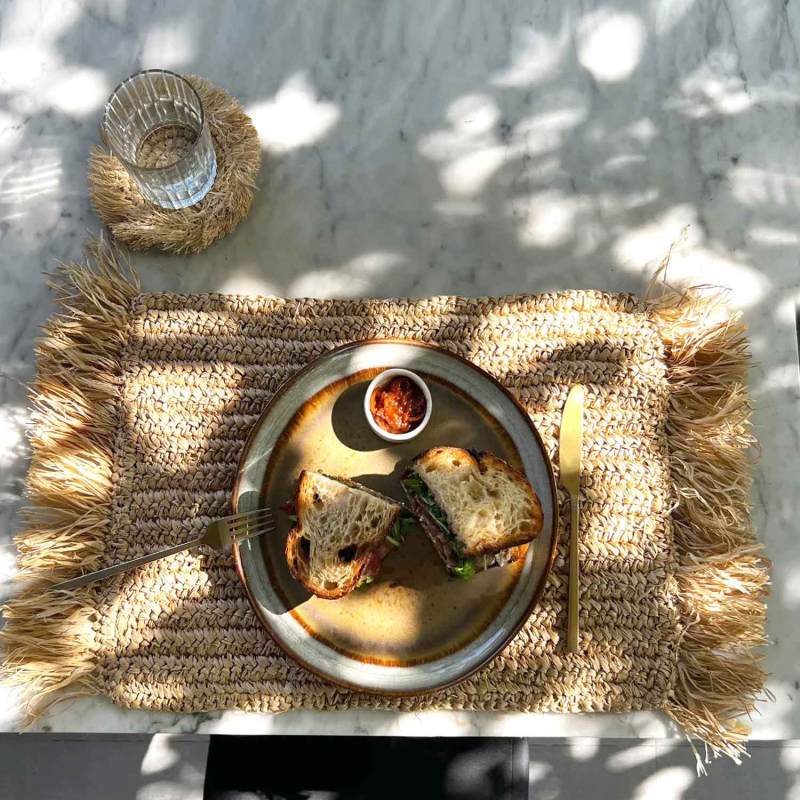 Preview: echteckiges Raffia Platzset mit Fransen auf Marmortisch, gedeckt mit Teller, Besteck und Glas im Sonnenlicht