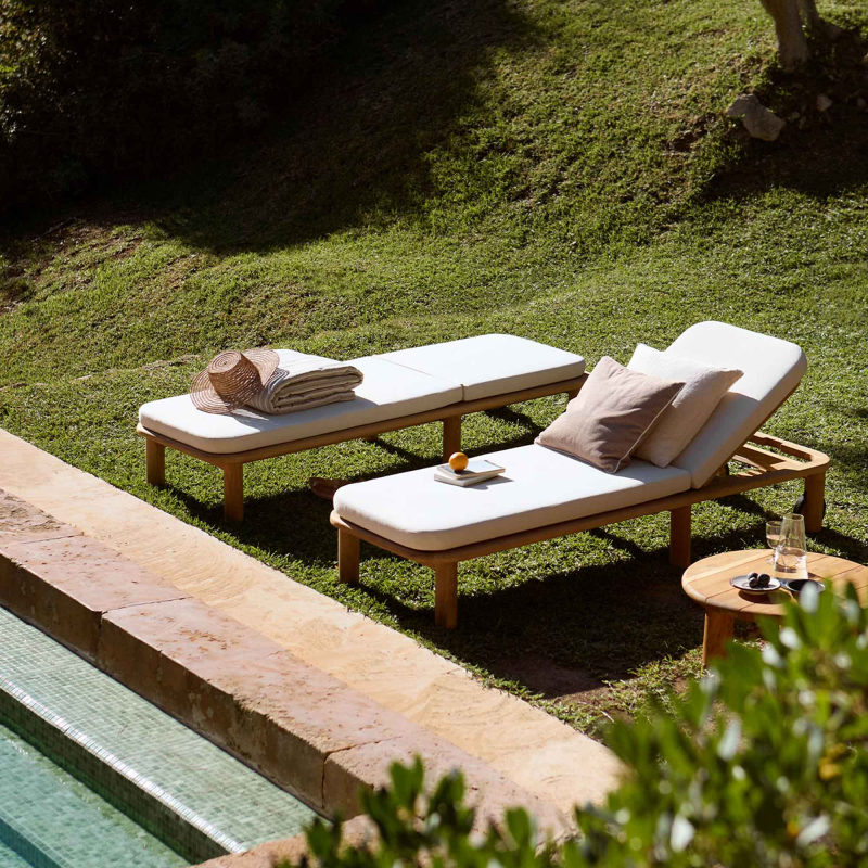 Zwei Tine K Teak Sonnenliegen am Pool — cremefarbene Kissen, Teak Beistelltisch, Steinmauer im Hintergrund