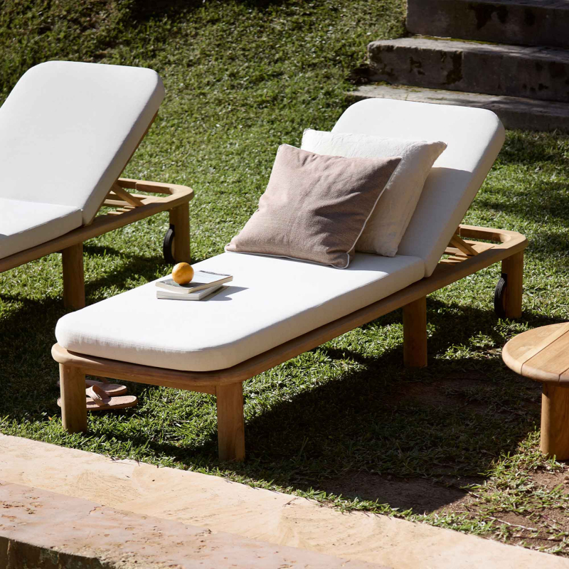 Preview: Tine K Teak Sonnenliege im Garten — cremefarbenes Quick-dry Kissen, verstellbare Rückenlehne, Kissen und Buch als Deko, sonniger Rasen