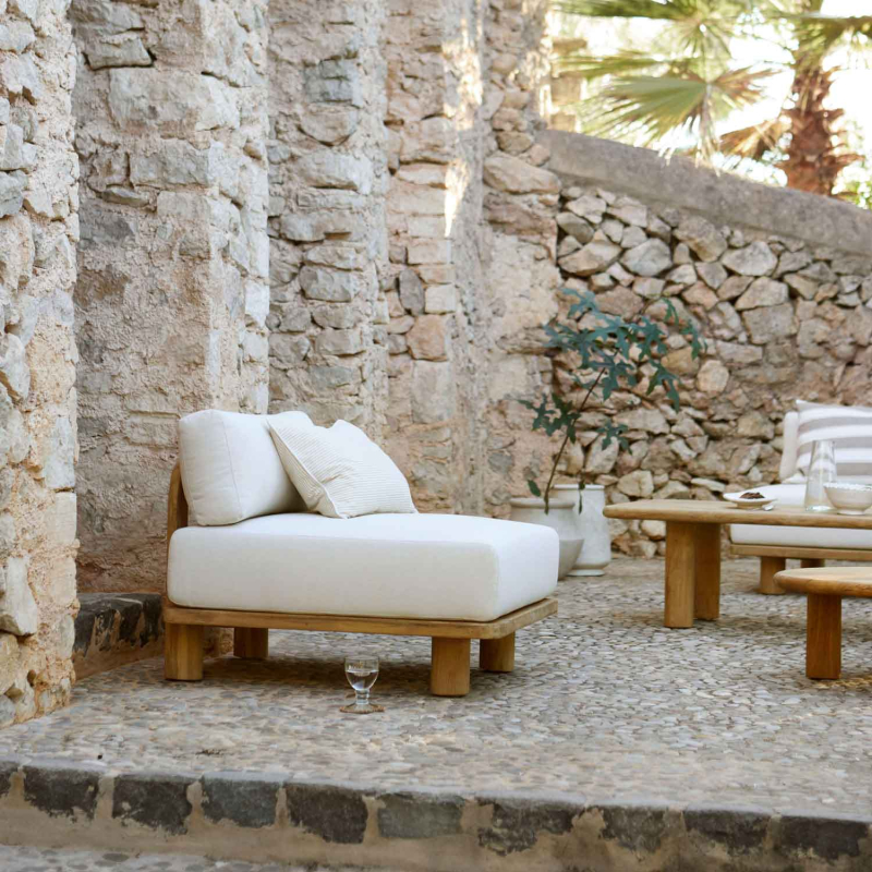 Tine K Teak Modul Sofa als Einzelsessel auf Kopfsteinpflaster — cremefarbene Kissen, recyceltes Teak, mediterraner Innenhof mit Steinmauer
