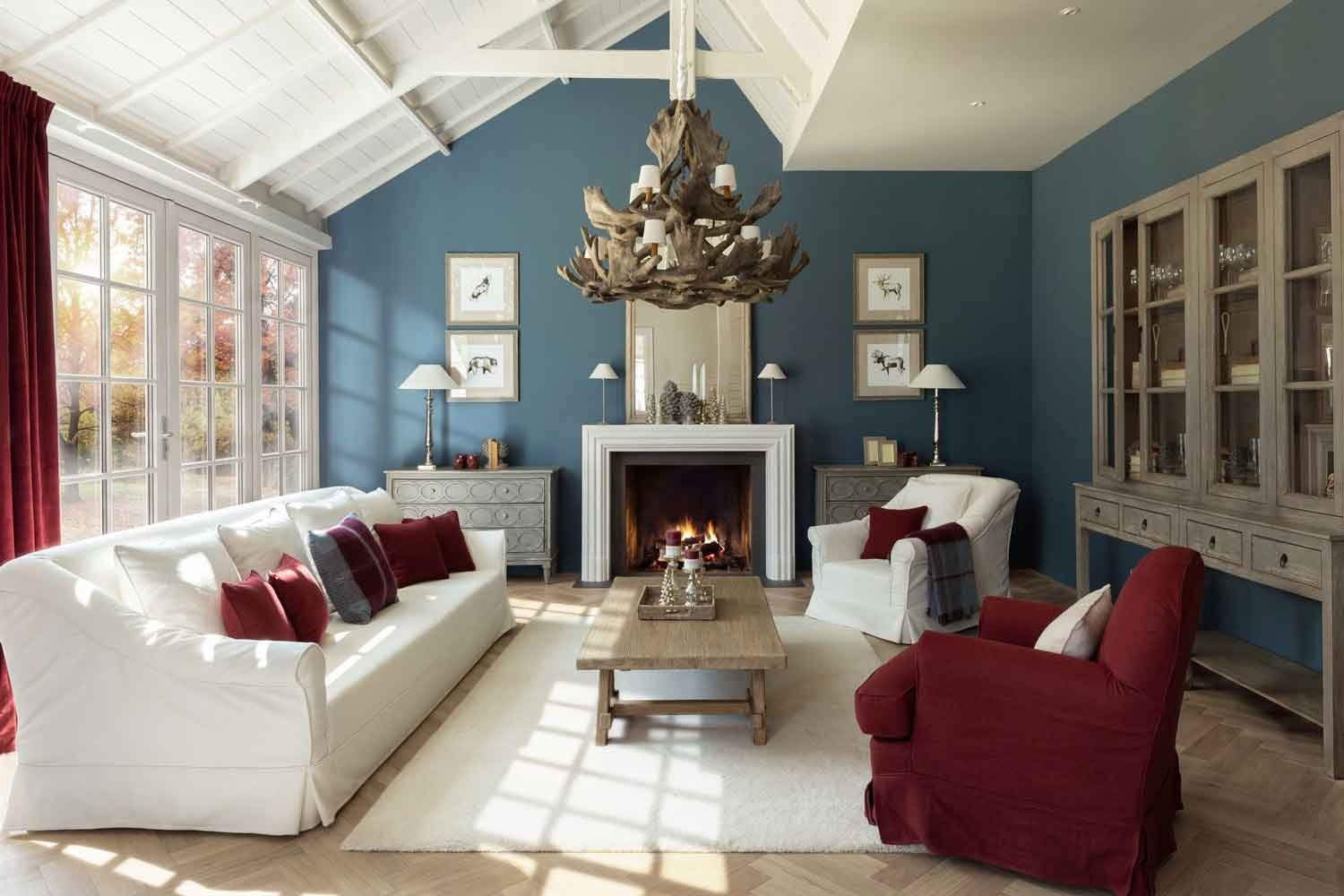 Wohnzimmer mit Kamin, weißem Sofa und Sesseln Bari, Kommoden Steeve, Couchtisch Benoit, Kronleuchter Sauvage und Vitrine an blauer Wand