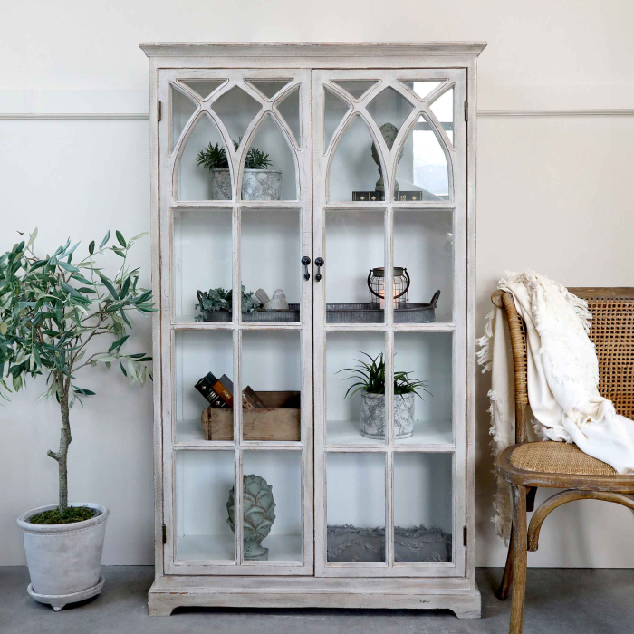 Vitrine mit gotischen Sprossenbogen-Glastüren im hellen Wohnzimmer — dekoriert mit Pflanzen und Vintage-Objekten, Provence-Stil